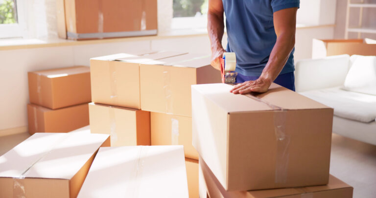 Person taping a cardboard moving box in a bright living room with stacked boxes around him/her.
